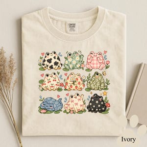 Peut inclure: Un t-shirt ivoire avec une impression graphique de neuf grenouilles stylisées disposées en grille. Les grenouilles présentent divers motifs dont des taches de vache, des fleurs, du vichy, des cerises, des fraises, des feuilles, des motifs cachemire, des nœuds et des étoiles.