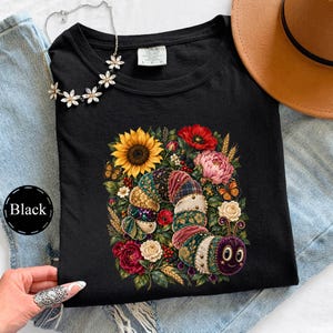 Könnte beinhalten: Ein schwarzes T-Shirt mit einer bunten Grafik einer segmentierten Raupe, umgeben von Sonnenblumen, roten Mohnblumen, rosa Pfingstrosen, weißen Rosen, Weizenähren und Schmetterlingen. Die Raupe hat einen violetten Kopf und gemusterte Körpersegmente.