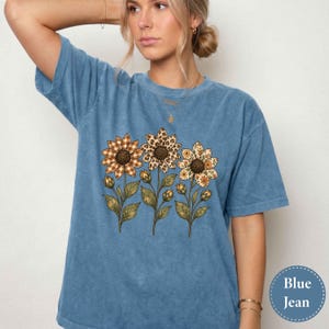 Könnte beinhalten: Ein blaues, mineralgewaschenes T-Shirt mit einem Grafikdruck von drei Sonnenblumen, deren Blütenblätter verschiedene Muster aufweisen, darunter ein braun-weißes Vichy-Karo, ein Leopardenmuster und ein Blumenmuster, mit grünen Stielen und Blättern.
