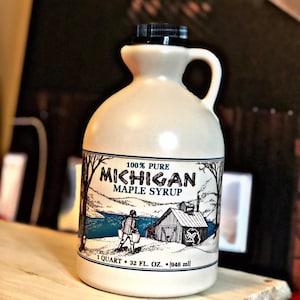 May include: A ceramic jug of Michigan maple syrup. The label reads "100% Pure Michigan Maple Syrup" with a scenic illustration. The jug holds 1 quart or 32 fl. oz. (946 ml) of syrup and has a black cap and handle.