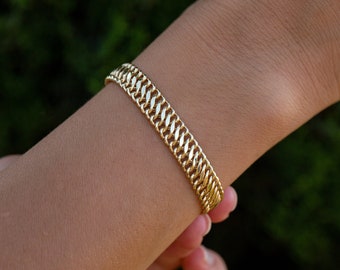 Könnte beinhalten: Ein goldfarbenes Armband mit Kettenglied-Design wird am Handgelenk getragen. Das Armband hat ein gewebtes Muster und besteht aus einem glänzenden, metallischen Material. Der Hintergrund ist ein verschwommenes Grün.