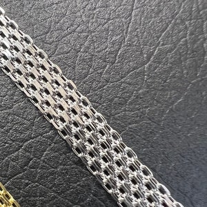 K&ouml;nnte beinhalten: Eine silberne Kettenkette mit einem zarten, verflochtenen Design. Die Kette liegt auf einer schwarzen, strukturierten Oberfl&auml;che.