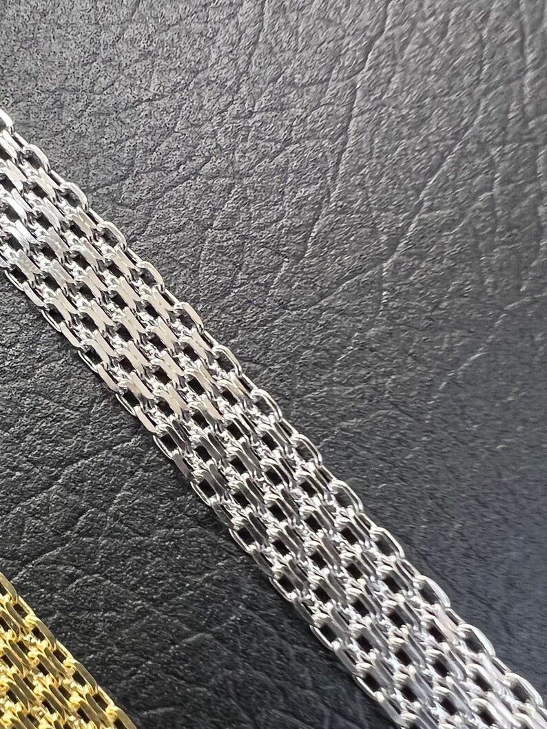 K&ouml;nnte beinhalten: Eine silberne Kettenkette mit einem zarten, verflochtenen Design. Die Kette liegt auf einer schwarzen, strukturierten Oberfl&auml;che.