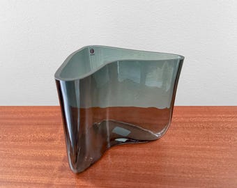 May include: A translucent, smoke-gray glass vase featuring a distinct, undulating, wave-like silhouette with curved, organic edges. The vessel has a smooth, reflective surface and a small, circular brand sticker visible on the upper rim.
