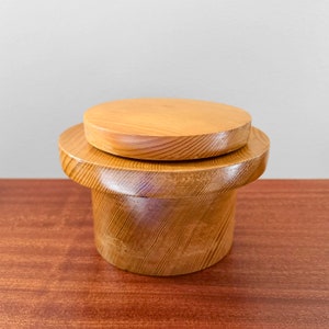 Op de afbeelding: Een houten container met een deksel. De container is gemaakt van lichtbruin hout en heeft een gladde, afgeronde vorm. Het deksel is ook van hout en past precies op de container.