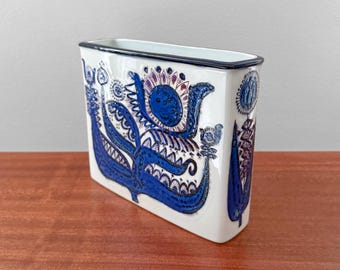 May include: A rectangular white ceramic vase featuring a dark blue and purple floral pattern. The design includes intricate stylized petals, leaves, and circular motifs across the front surface. The vase has a thin dark blue rim along the top edge.