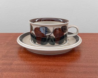 May include: A ceramic teacup and saucer set. The cup and saucer are beige with a brown and gray rim. The cup features a pattern of brown and green leaves. The set is placed on a wooden surface.