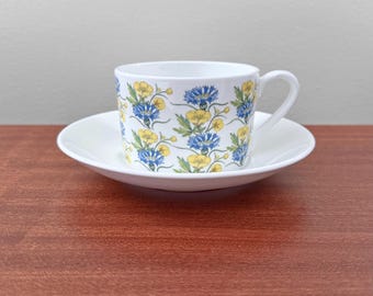 May include: White teacup and saucer set with a floral pattern. The cup and saucer are white with a repeating pattern of blue and yellow flowers with green leaves. The cup has a curved handle.
