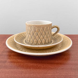 May include: A ceramic cup and saucer set with a matching plate, featuring a tan and brown leaf-like embossed pattern. The cup has a curved handle and a white interior, while the saucer and plate display a radiating textured design in earthy tones.