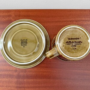 May include: Overhead shot of a vintage Arabia Kosmos ceramic cup and saucer set. The set is olive green with a brown rim and features the Arabia logo and "Kosmos" text. The saucer is 6 inches in diameter.
