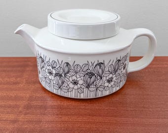 May include: White ceramic teapot with a lid, featuring a black and white floral pattern around the base. The teapot has a curved handle and a spout for pouring. The lid has a black trim.