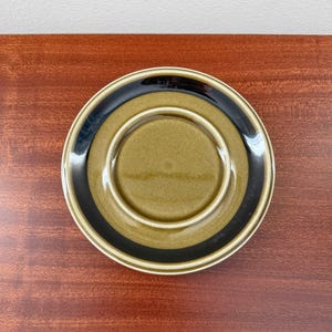May include: A ceramic saucer with a circular design. The saucer features a central olive-green area, a black band, and a cream-colored outer rim. The saucer is set against a dark wood surface.