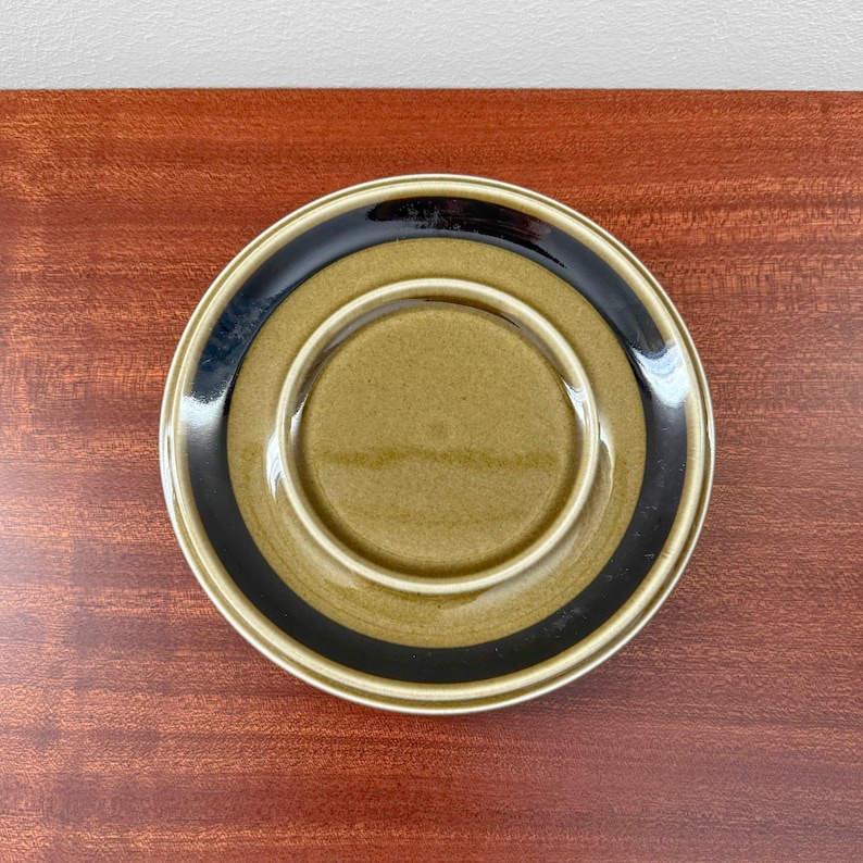 May include: A ceramic saucer with a circular design. The saucer features a central olive-green area, a black band, and a cream-colored outer rim. The saucer is set against a dark wood surface.