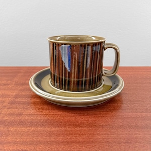 Peut inclure: Un ensemble tasse et soucoupe à café en céramique marron et beige. La tasse présente des rayures verticales dans les tons marron et beige. La soucoupe a une bordure bleu foncé et beige. L'ensemble est posé sur une surface en bois.