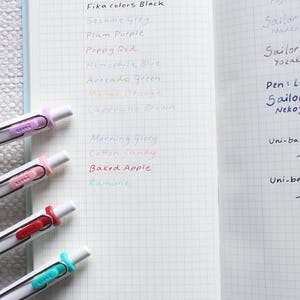 May include: An open notebook with squared paper, displaying pen tests. The left page lists pen colours like Poppy Red and Avocado Green. The right page shows pen brands such as Sailor Shikiori and Lamy Safari. Several pens with white barrels and coloured accents are visible.