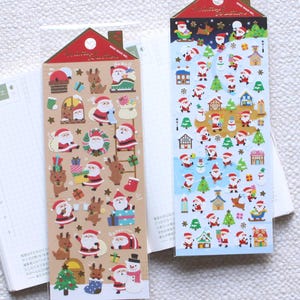 Puede incluir: Dos hojas de pegatinas con la parte superior en forma de casa, con ilustraciones navideñas festivas. Una hoja tiene a Papá Noel, renos y regalos sobre un fondo beige. La otra tiene a Papá Noel, árboles y casas sobre un fondo azul.