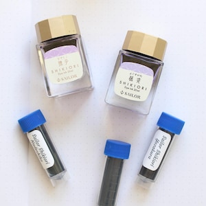 An overhead shot of Sailor Shikiori dye ink bottles and vials. Two square glass bottles with gold lids and labels that read "SHIKIORI Dye ink 20ml" are visible. Three vials with blue caps are also present, labeled "Sailor Shikiori".