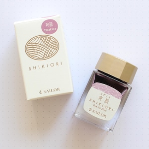 A clear glass bottle of Shikiori dye ink, 20ml, with a gold cap and a label featuring Japanese text and a pink design. Beside it is a white box with the Shikiori logo and text, and the Sailor logo.