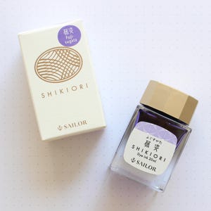 A clear glass bottle of Shikiori dye ink, 20ml, with a gold cap and a white label with Japanese text. The ink is a purple color. A matching box with the text "SHIKIORI" and "SAILOR" is next to the bottle.