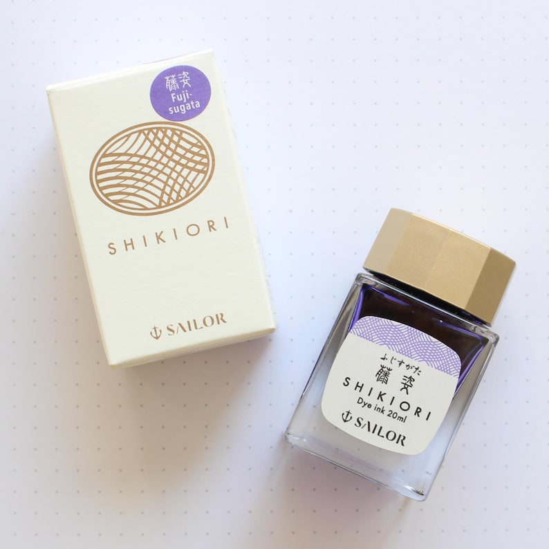 A clear glass bottle of Shikiori dye ink, 20ml, with a gold cap and a white label with Japanese text. The ink is a purple color. A matching box with the text "SHIKIORI" and "SAILOR" is next to the bottle.