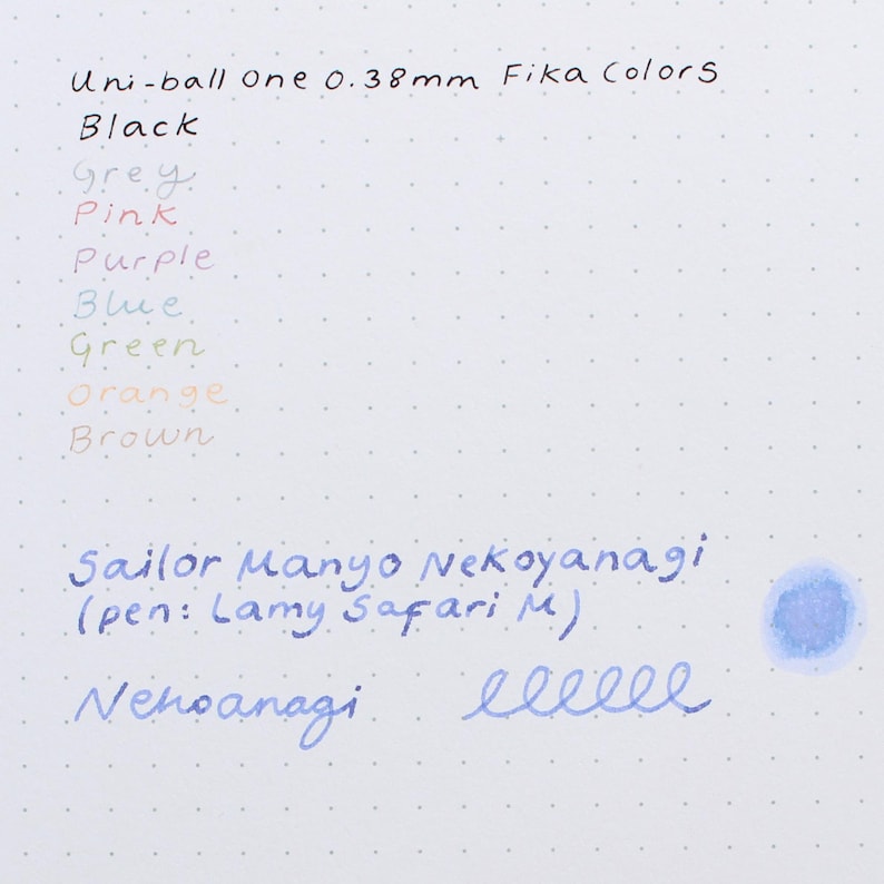 A close-up of a white paper with handwritten ink swatches in various colors. The text includes "Uni-ball One 0.38mm Fika Colors" and "Sailor Manyo Nekoyanagi". Colors include black, grey, pink, purple, blue, green, orange, and brown.