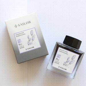 A clear glass ink bottle and matching box. The bottle and box have labels with the text "MANYO INK NEKOYANAGI" and "SAILOR". The labels also feature a floral illustration and state the ink volume is 50ml. The box is gray and white.