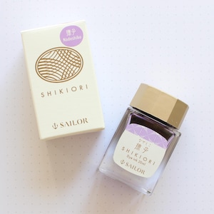 A clear glass bottle of Shikiori dye ink, 20ml, with a gold cap and a white label with Japanese text. The ink is a light purple color. A matching box with the text "SHIKIORI" and "SAILOR" is next to the bottle.