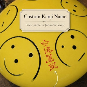 May include: A bright yellow steel tongue drum featuring multiple circular sound cutouts and a vertical column of red Japanese kanji characters. A white rectangular label with the text Custom Kanji Name and Your name in Japanese kanji is visible.