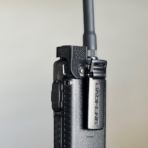 Puede incluir: Una radio bidireccional de mano negra con una antena negra y un clip de cinturón negro. La radio tiene el texto "BAOFENG" impreso en el lateral.