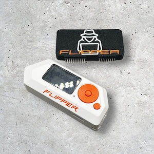 May include: A white and orange Flipper device with a small screen and buttons. The device is on a grey concrete surface. A black rectangular device with the word "Flipper" in orange letters is also on the surface.