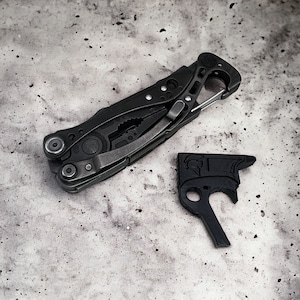 May include: A black multi-tool with a variety of attachments, including a pliers, a knife, and a screwdriver. The tool is lying on a gray textured surface. A black plastic object with a hole in the center is also on the surface.