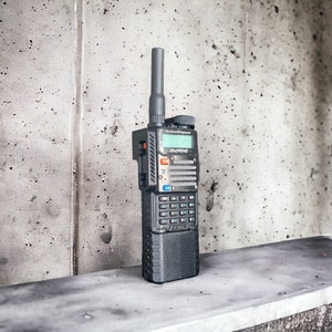 May include: A black handheld two-way radio with a silver antenna and a black and silver keypad. The radio has a small LCD screen with the text "Dual Band VHF/UHF Transceiver" and the brand name "BAOFENG" displayed.