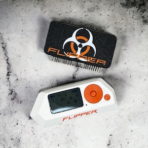 May include: A white and orange electronic device with a screen displaying icons and the word "FLIPPER" printed on the front. A black sticker with a biohazard symbol and the word "FLIPPER" is also shown.