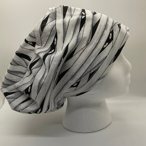 May include: A white and black patterned scrub hat with a mummy design. The hat has black eyes and a white background with black stripes.
