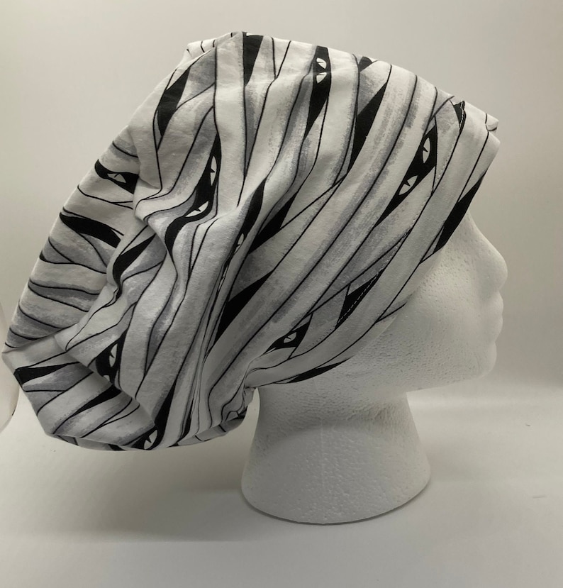 May include: A white and black patterned scrub hat with a mummy design. The hat has black eyes and a white background with black stripes.