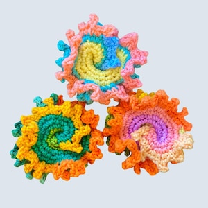 Könnte beinhalten: Drei handgefertigte Häkelspiralen in leuchtenden Farben. Jede Spirale zeigt eine einzigartige Mischung aus hellen Farbtönen, darunter Gelb, Orange, Rosa, Blau und Grün. Die Kanten sind gewellt und verleihen dem Kunsthandwerk ein strukturiertes, dekoratives Element.
