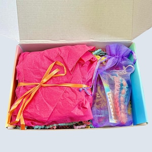 May include: An open cardboard box filled with colorful items. A bright pink tissue paper package tied with an orange ribbon sits next to a purple mesh bag. The box is lined with a rainbow gradient.