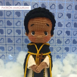 Puede incluir: Muñeco amigurumi de crochet de San Martín de Porres, un santo de la Iglesia Católica. El muñeco lleva una túnica negra y blanca con ribetes dorados y sostiene una cruz. El muñeco tiene piel morena y cabello negro. El fondo es un patrón azul y blanco de corazones.