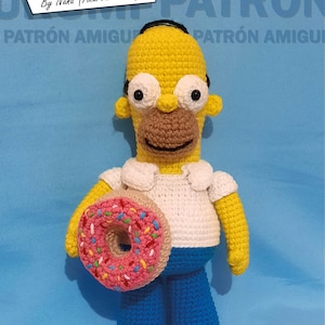 Puede incluir: Un amigurumi de Homer Simpson tejido a crochet sosteniendo una dona rosa con chispas de colores. La muñeca lleva una camisa blanca y pantalones azules. El fondo es una superficie azul claro con el texto "Homero By Nana (Andrea Sierra)" y "PATRÓN AMIGURUMI" en estilo de cómic.