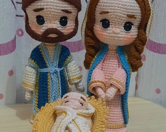 Puede incluir: Escena de la natividad en amigurumi de crochet con un José de cabello castaño, una María vestida de rosa y un bebé Jesús en un pesebre amarillo. El título "Nacimiento by Nuna (Andrea Sierra)" está escrito debajo de las figuras.