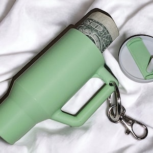 May include: A light green silicone keychain with a small, removable container. The container is open and a dollar bill is partially visible inside. The keychain has a silver metal clasp.