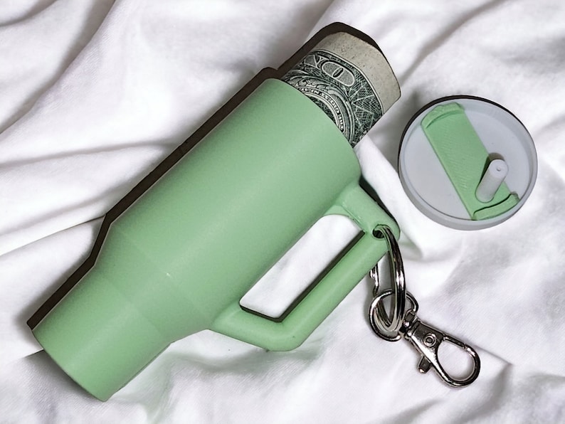 May include: A light green silicone keychain with a small, removable container. The container is open and a dollar bill is partially visible inside. The keychain has a silver metal clasp.