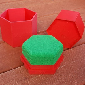 May include: Three red hexagonal boxes, one with a green hexagonal lid. The boxes are made of plastic and have a smooth surface. The boxes are stacked on top of each other.