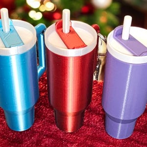May include: Three travel mugs with lids and straws. The mugs are blue, red, and purple. The mugs have a handle and a keychain.