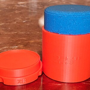 May include: A red plastic container with a blue lid. The container has the text "2.5" printed on the side. The lid is slightly smaller than the container and has a textured surface.