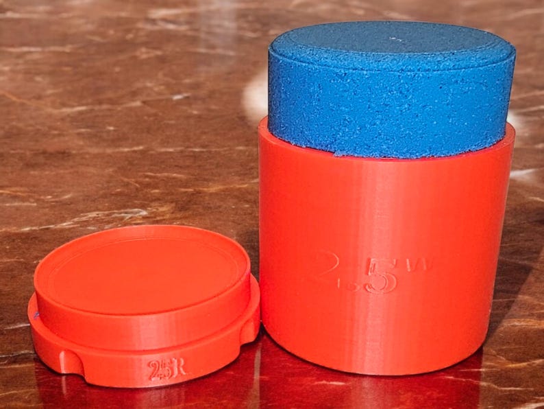 May include: A red plastic container with a blue lid. The container has the text "2.5" printed on the side. The lid is slightly smaller than the container and has a textured surface.