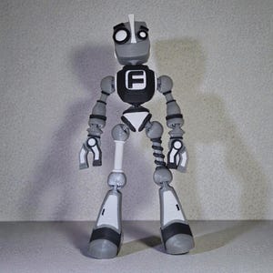 May include: A gray and white articulated robot figurine with a black chest emblem featuring a white "F". The robot has a cylindrical head, jointed limbs, and a white and black color scheme. The robot's design is reminiscent of a vintage toy.
