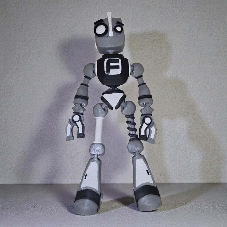 May include: A gray and white articulated robot figurine with a black chest emblem featuring a white "F". The robot has a cylindrical head, jointed limbs, and a white and black color scheme. The robot's design is reminiscent of a vintage toy.