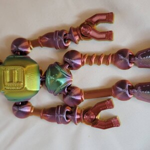 May include: A multi-colored, articulated robot toy with a metallic finish. The toy features a head, torso, arms, and legs, all connected by joints. The color scheme includes shades of gold, green, and purple.