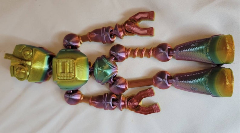 May include: A multi-colored, articulated robot toy with a metallic finish. The toy features a head, torso, arms, and legs, all connected by joints. The color scheme includes shades of gold, green, and purple.